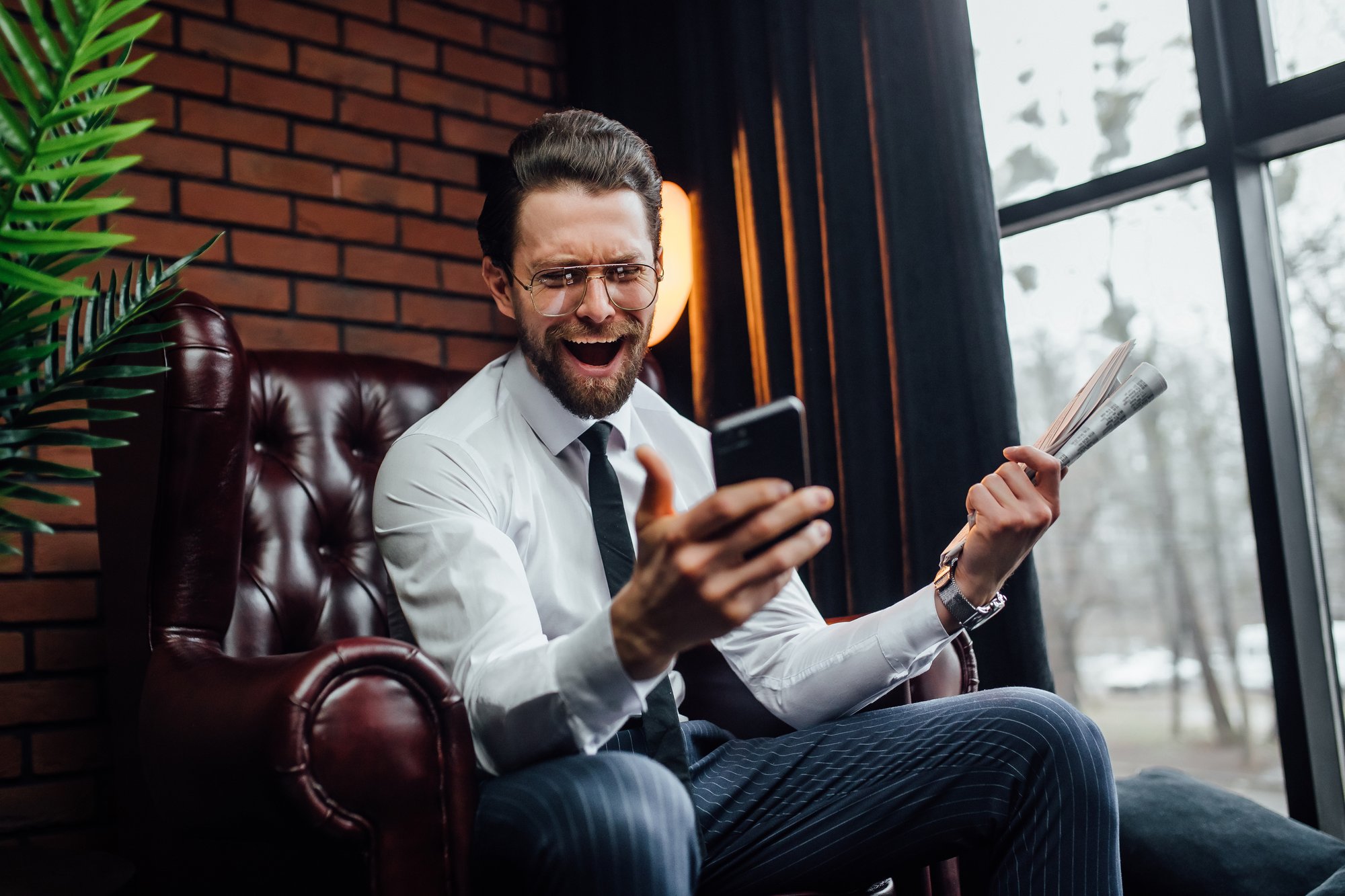 handsome-businessman-with-candid-emotions-holding-his-smartphone-newspaper-while-sitting-armchair